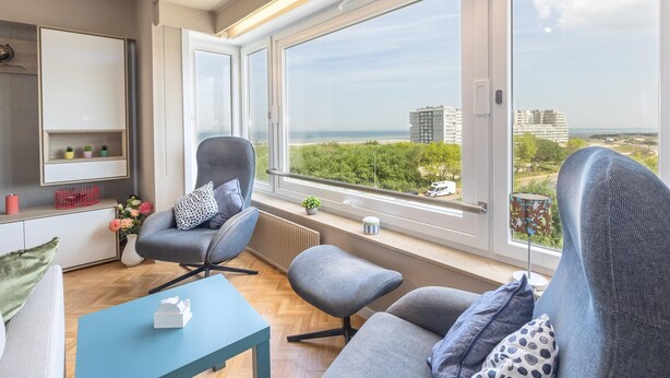 Un salon moderne avec une grande fenêtre, un canapé gris, une petite table et un radiateur.