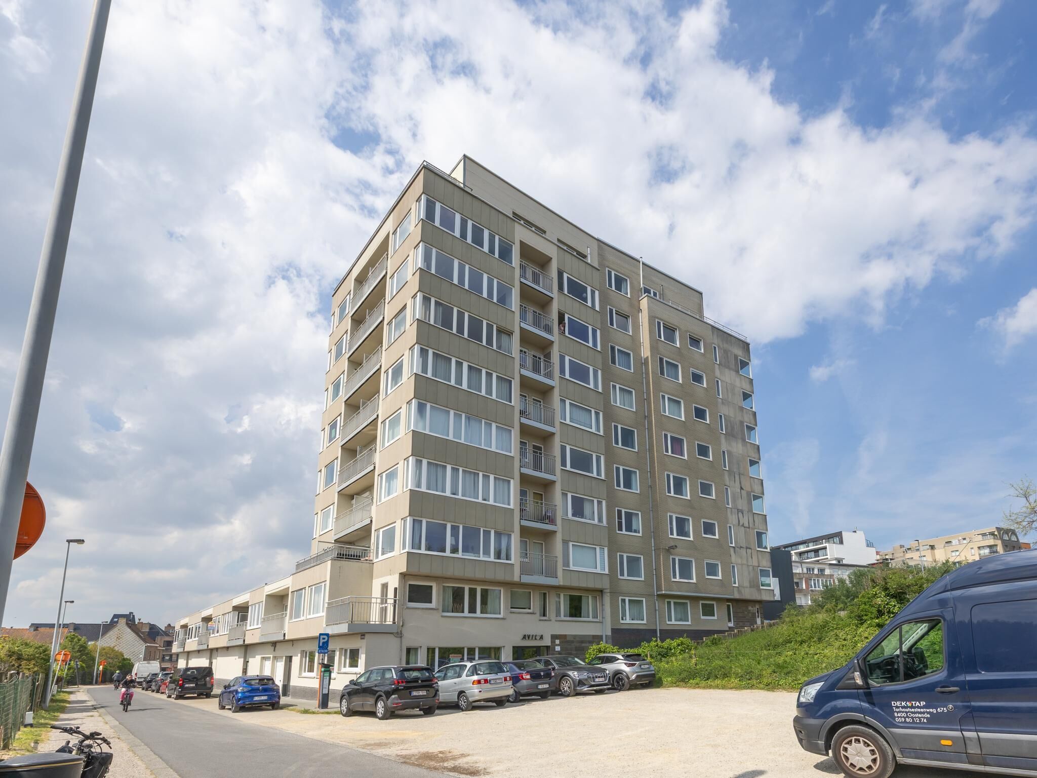 A multi-story building with a parking area in front, a blue van parked to the side, and a clear sky with clouds.