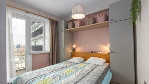 Une chambre avec un lit, une fenêtre, une plante suspendue et une étagère avec des sacs.