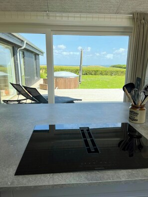 A modern kitchen with a large black induction cooktop, a glass door leading to a balcony with a view of the sea, and a wooden hot tub. - Summer house with wilderness bath and water views (Vinderup)