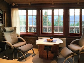 Ein gemütliches Zimmer mit einem Holztisch, zwei Stühlen und großen Fenstern mit Blick auf Bäume und Berge.