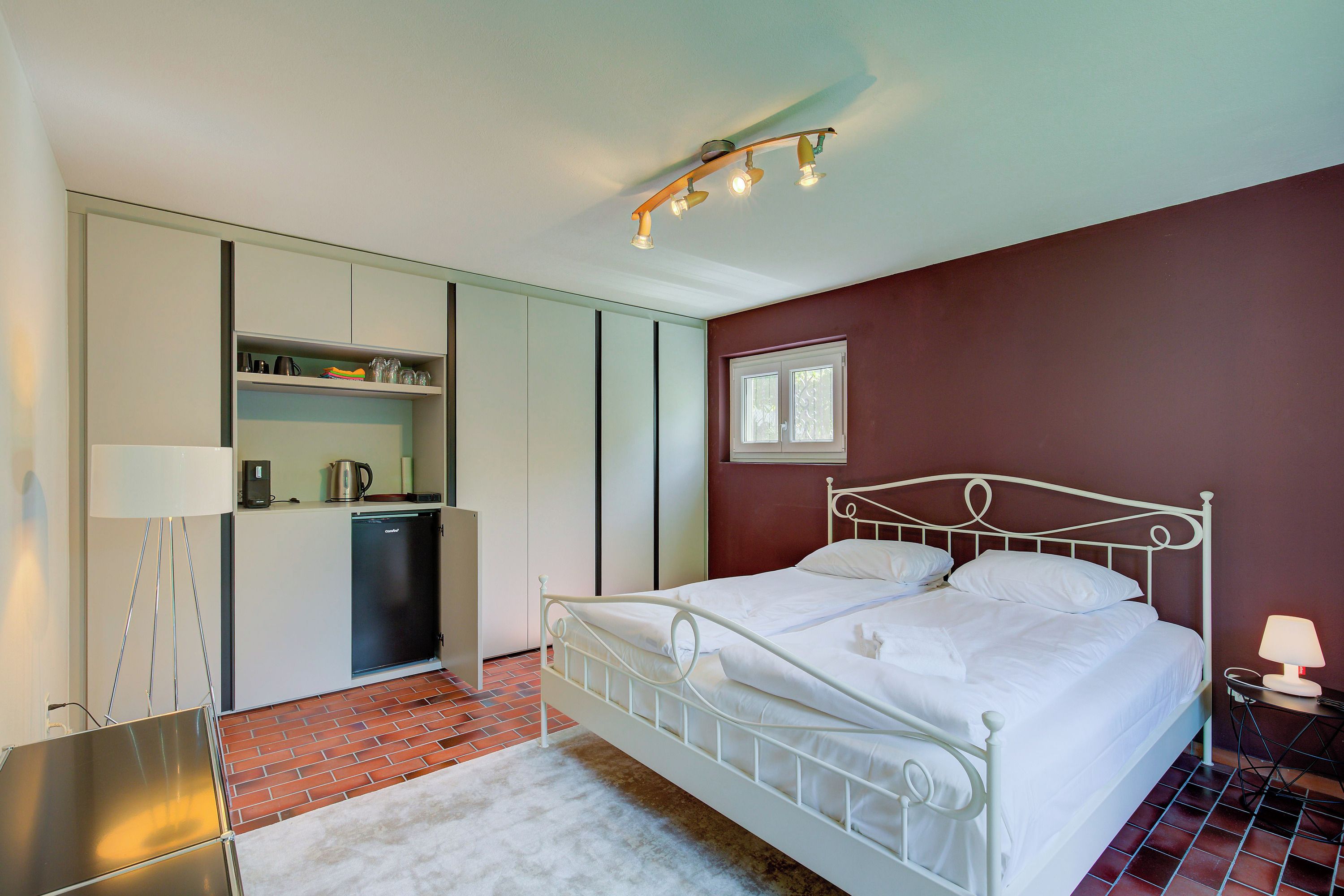 A bedroom with a white metal bed, a small bedside table with a lamp, a kitchen area with a refrigerator, and a window with blinds.