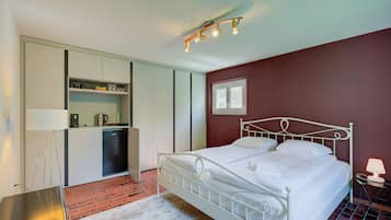 A bedroom with a white metal bed, a small bedside table with a lamp, a kitchen area with a refrigerator, and a window with blinds.