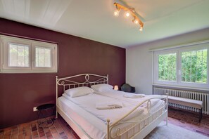 Un dormitorio con cama, un banco, un radiador y una ventana con vistas al verdor.