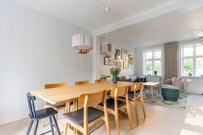 A modern dining room with a wooden table and chairs, a hanging light fixture, and a sofa in the background.