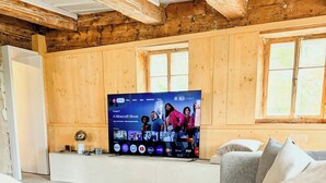 Um quarto aconchegante com uma cama, uma televisão exibindo um filme, paredes de madeira e uma janela.