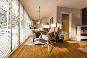 Ein modernes Wohnzimmer mit Essbereich, Holzboden, Kamin und großen Fenstern mit Jalousien.