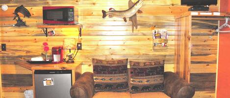 A compact room with a wooden wall, a small kitchenette, a sofa with patterned cushions, and a mounted television.