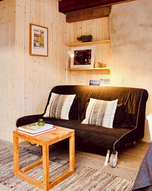 A cozy living room with a wooden floor, a black sofa with patterned cushions, a wooden coffee table with books, and a framed poster on the wall.