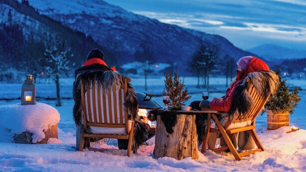 Dos personas sentadas en una mesa de madera al aire libre, entre la nieve, con un farol y un pequeño árbol sobre la mesa.