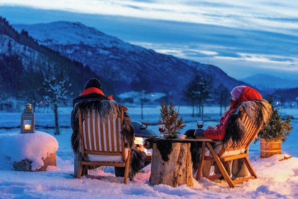 Zwei Personen sitzen draußen an einem Holztisch im Schnee, mit einer Laterne und einem kleinen Baum darauf.