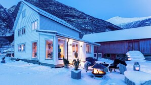 Una casa de dos pisos con techo nevado, un porche con una chimenea y montañas al fondo.