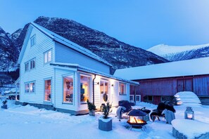 Ein zweistöckiges Haus mit verschneitem Dach, einer Veranda mit Feuerstelle und einem Berg im Hintergrund.