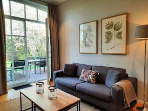 Ein Wohnzimmer mit grauem Sofa, zwei gerahmten Pflanzenbildern, einem Couchtisch mit Kerzen und Blick auf eine Terrassenanlage.