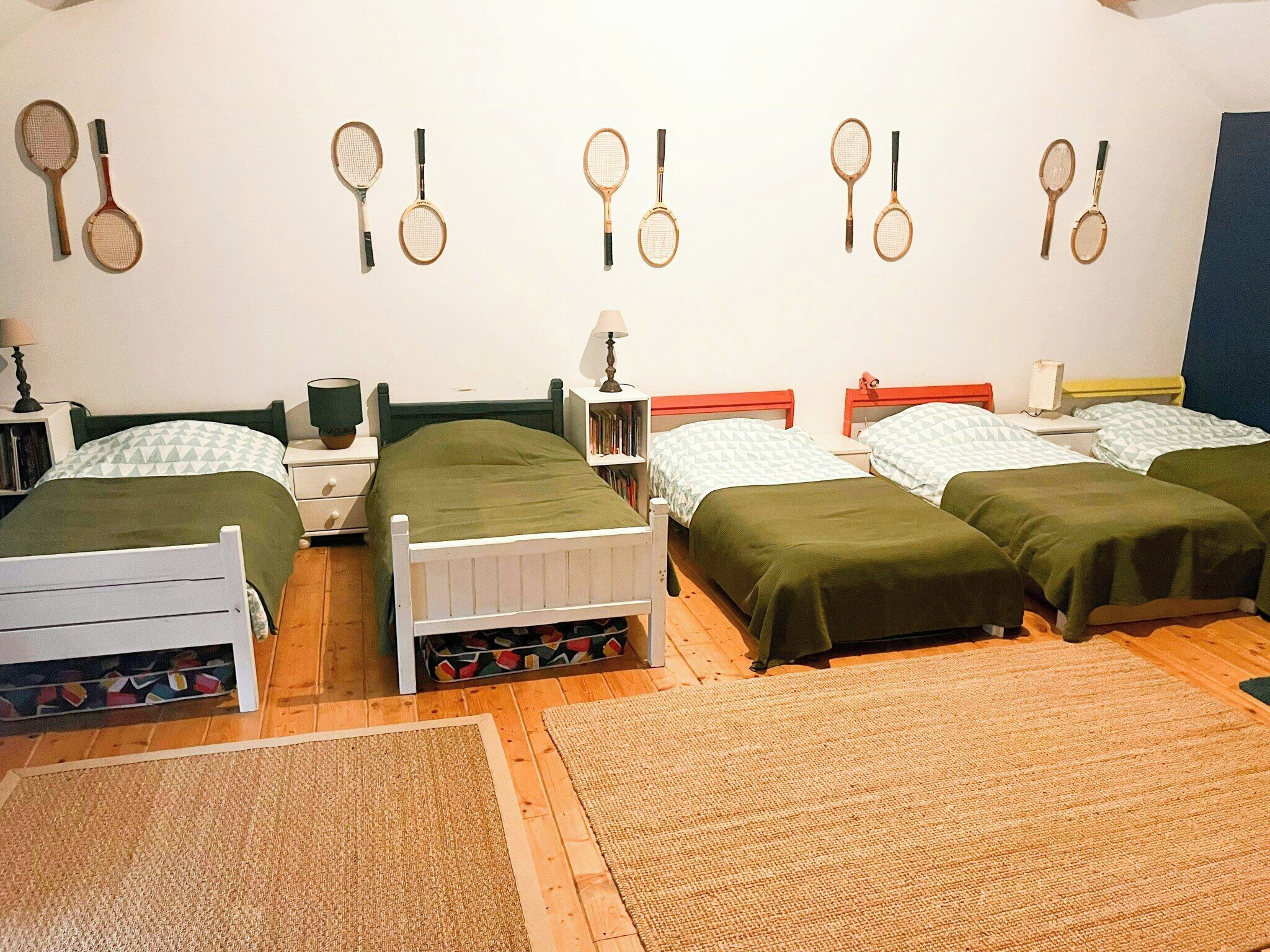 A room with four single beds, a wall-mounted rack of tennis rackets, a bedside table with a lamp, and a small bookshelf.