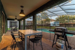 Eine Terrasse im Freien mit Holztisch und -stühlen, einem Grill und einem Swimmingpool im Hintergrund.