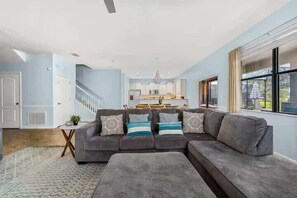 Ein geräumiges Wohnzimmer mit einem grauen Ecksofa, einem kleinen Beistelltisch und einer Treppe, die zu einer oberen Ebene führt.