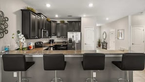 A modern kitchen with dark cabinets, stainless steel appliances, and a granite countertop. There are three black bar stools in front of the counter. - 9BR Themed Close to Disney World! Movie Theatre! (Four Corners)