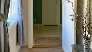 Um corredor com porta, tapete de entrada, planta em vaso e janela com cortinas.