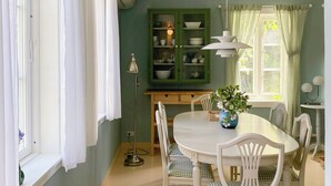 Uma sala de jantar com uma mesa e cadeiras brancas, um armário verde com louças e uma janela com cortinas.