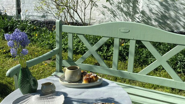 Um banco de madeira verde com uma mesa branca à frente, um vaso com flores roxas e um prato com comida e uma xícara.
