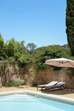 Ein Swimmingpool mit Liegestühlen und einem Sonnenschirm, umgeben von einer Steinmauer und Grünpflanzen.
