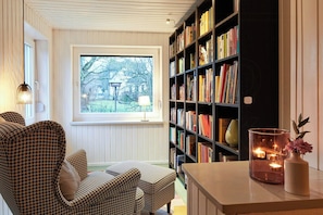 A cozy reading nook with a houndstooth armchair, a bookshelf filled with books, and a window with a view of trees. - Holiday home with garden and sauna in Mecklenburg (Hohen Demzim)