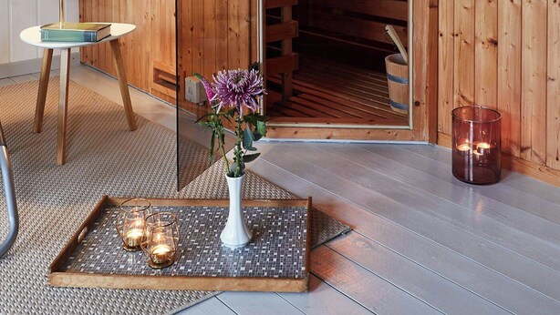 Un sauna en bois avec une porte en verre, un porte-serviettes, une petite table avec une lampe, et un plateau avec des bougies et un vase.