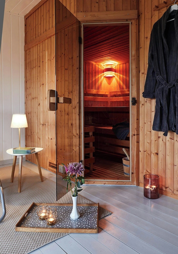 Een houten sauna met een glazen deur, een handdoekrek, een klein tafeltje met een lamp, en een dienblad met kaarsen en een vaas.