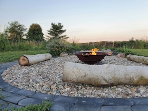A circular fire pit with logs, surrounded by pebbles, set in a garden with trees and a clear sky. - Holiday home with garden and sauna in Mecklenburg (Hohen Demzim)