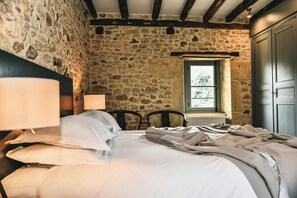 Une chambre avec des murs en pierre, un plafond en bois, un lit recouvert de draps blancs, une fenêtre et une porte.