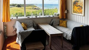 A cozy room with a window view of a grassy area and the sea, a wooden table, two sofas, and a framed artwork on the wall. - Wooden house with panoramic view over Kattegat (Hundested)
