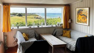 Et hyggeligt værelse med udsigt til græsareal og havet, et træbord, to sofaer og et billede i ramme på væggen.