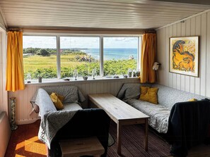 Ein gemütliches Zimmer mit Blick aus dem Fenster auf eine grüne Fläche und das Meer, ein Holztisch, zwei Sofas und ein gerahmtes Kunstwerk an der Wand.