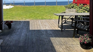 A wooden deck with a view of the ocean, a red building, and a grassy area. - Wooden house with panoramic view over Kattegat (Hundested)
