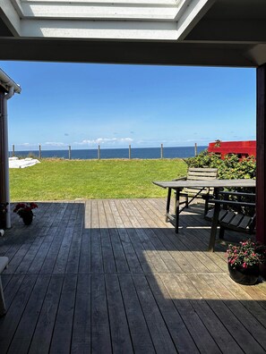 Eine Holzterrasse mit Blick auf das Meer, ein rotes Gebäude und eine Rasenfläche.