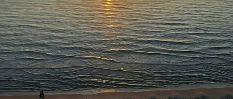 A sunset over a beach with calm water, a person walking, and scattered rocks.