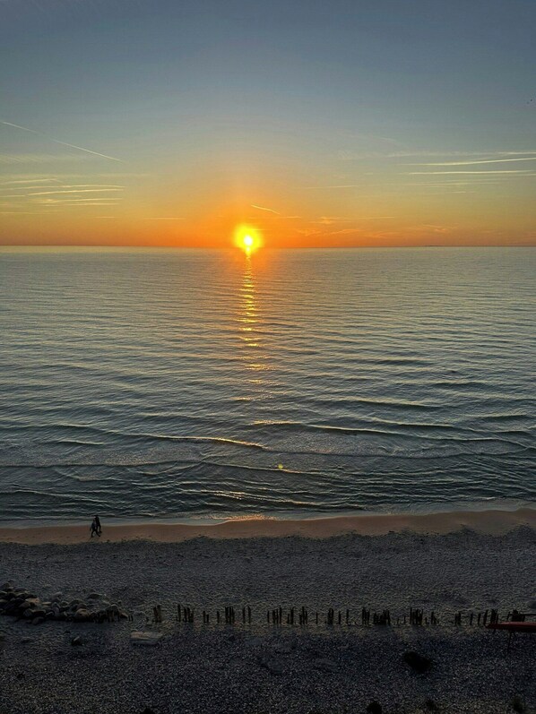 Ein Sonnenuntergang über einem Strand mit ruhigem Wasser, eine Person geht spazieren, und verstreute Felsen.