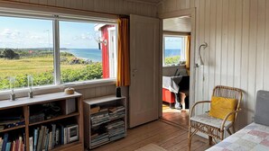 Et værelse med trægulv, en seng med et patchworktæppe, en fletstol med en gul pude, en reol med bøger og en lampe, samt havudsigt gennem et vindue.