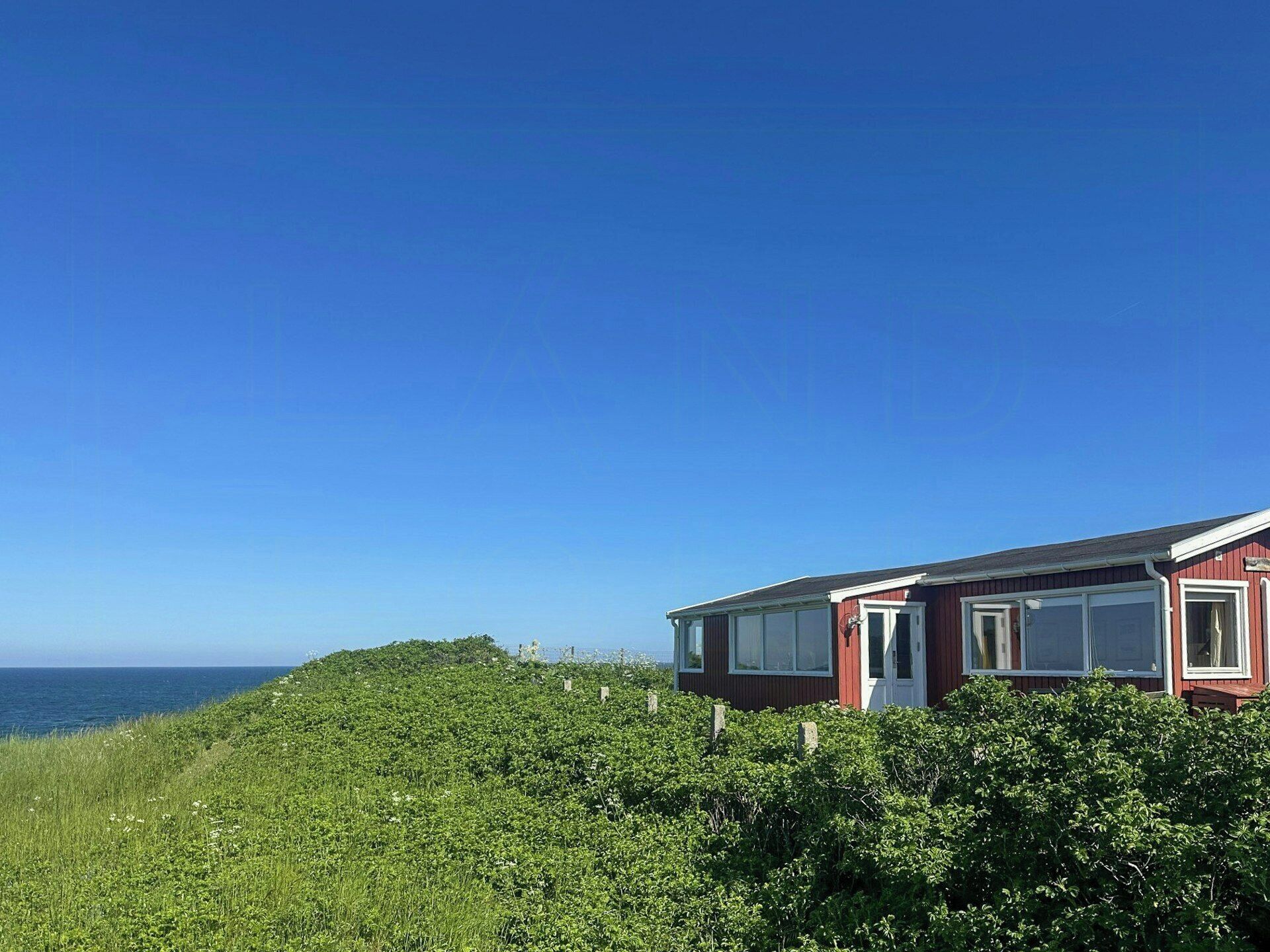 Et rødt hus med en hvid dør og vinduer, omgivet af grønt og med udsigt over havet.
