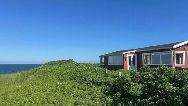 Et rødt hus med en hvid dør og vinduer, omgivet af grønt og med udsigt over havet.
