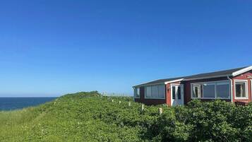 Et rødt hus med en hvid dør og vinduer, omgivet af grønt og med udsigt over havet.