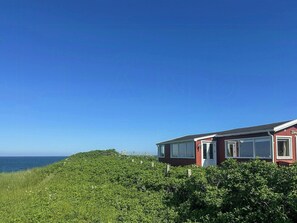 Ein rotes Haus mit weißer Tür und Fenstern, umgeben von viel Grün und mit Blick auf das Meer.