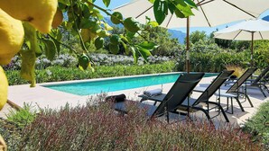 A poolside area with lounge chairs and umbrellas, surrounded by greenery and a clear sky. - 1700s design stone house in Mallorca with pool (Soller)