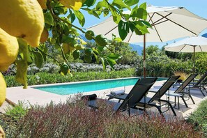 A poolside area with lounge chairs and umbrellas, surrounded by greenery and a clear sky. - 1700s design stone house in Mallorca with pool (Soller)