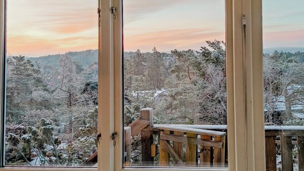 En vy genom ett fönster med en träveranda och snötäckta träd.