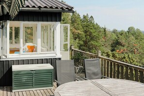 Eine Holzterrasse mit Tisch und Stühlen, ein Fenster mit Vorhängen und Blick auf Bäume.