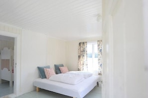 Ein Schlafzimmer mit Bett, Kissen, einem Fenster mit Vorhängen und einer Tür zu einem weiteren Raum.