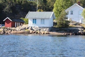 A small white building with a metal roof, a red shed, and a white house in the background, all situated by a body of water.