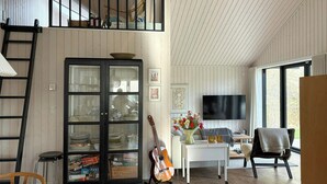 A loft-style living space with a loft bed, a glass-fronted cabinet with dishes, a guitar, a TV, and a view of the outdoors. - Cozy new vacation home near the water (Tisvilde)